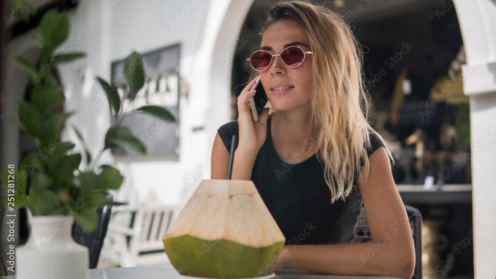 Portrait of happy beautiful tanned female in cafe in open air, drink in a coconut on the table, girl chatting with her friends by modern phone, wearing fashionable sunglasses, copy space
