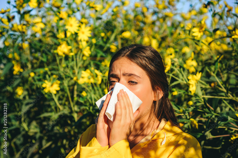 Sneezing young girl with nose wiper among blooming flowers in park. Young woman got nose allergy, flu sneezing nose. The girl suffers from pollen allergy during flowering and uses napkins. Allergy. Sneezing young girl with nose wiper among blooming flowers in park. Young woman got nose allergy, flu sneezing nose. The girl suffers from pollen allergy during flowering and uses napkins. Allergy.