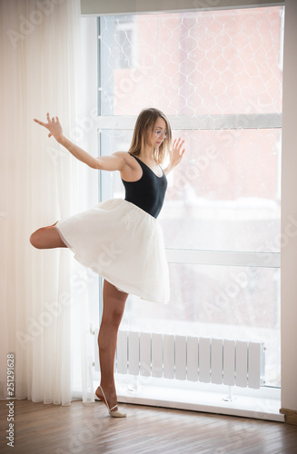 Fototapeta Naklejka Na Ścianę i Meble -  Ballerina girl in glasses stands on one leg near the window, in the afternoon in a bright studio