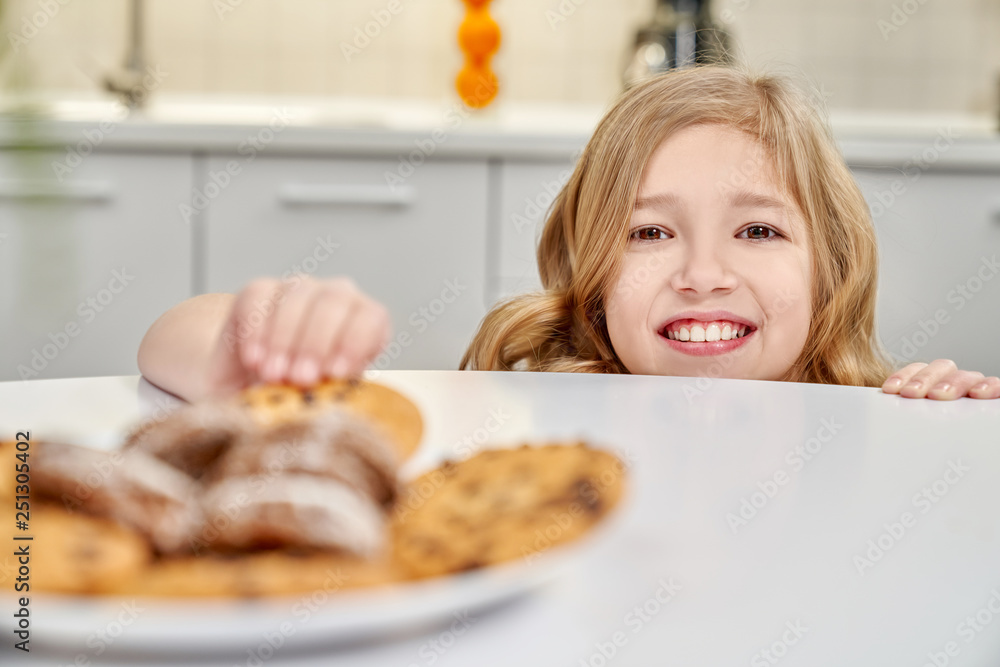 Child hiding and taking biscuits with raisins from table.