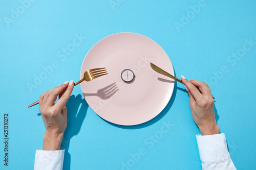 White plate with round whatch shows six o'clock served knife and fork in a girl's hands on a blue background. Time to eat and diet concept. Top view. photo