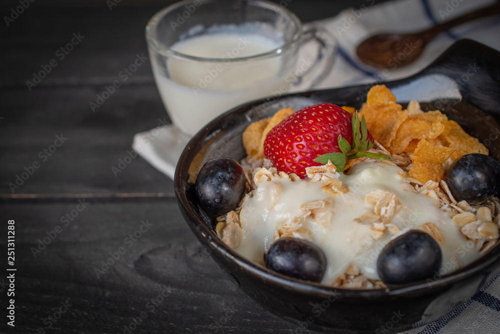 Yoghurt mix oatmeal, strawberry and grape topping in black bowl on Blue and white striped fabric and wood table with spoon and milk in glass placed back side. Yoghurt mix oatmeal, strawberry and grape topping in black bowl on Blue and white striped fabric and wood table with spoon and milk in glass placed back side.