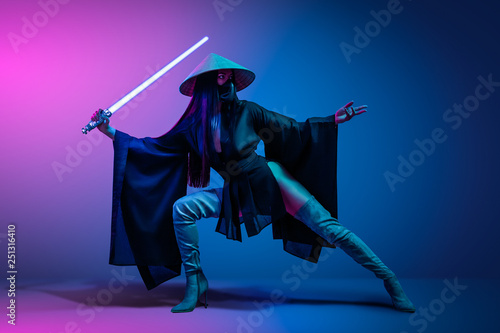 Concept on cosmic cosplay. Сontemporary portrait a young athletic woman in traditional Japanese black kimono, an Asian hat and highboots is holding a lightsaber and posing on neon blue-pink background photo