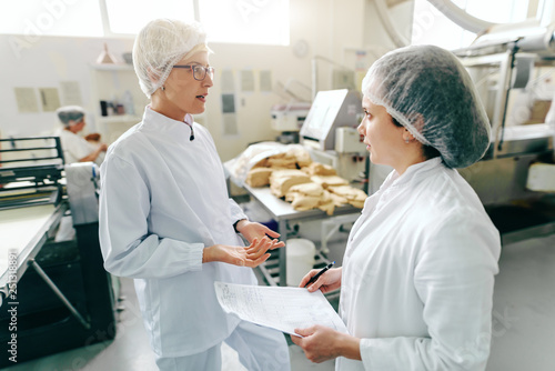 Two female caucasian employees talking about work while standing in food factory. One of them holding paperwork. In background machines.