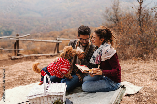 Smiling happy couple having date in nature. Man holding sandwich and petting poodle while woman reading a book. Autumn time.