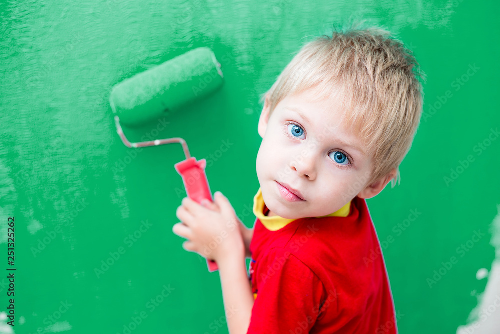 Cute blue-eyed blond pretty boy of five years in the red t-shirt makes repair paints the wall in blue roller