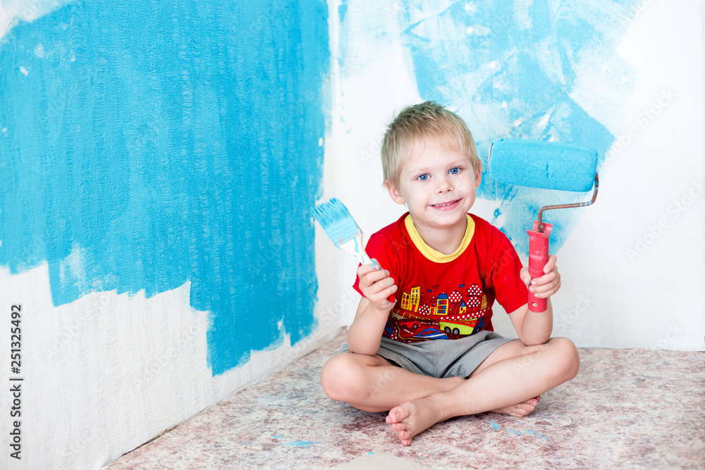 Fototapeta premium Cute blue-eyed blond pretty boy of five years in the red t-shirt makes repair paints the wall in blue roller