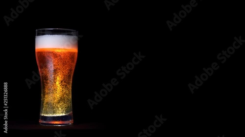 Glass of golden craft beer with white foam and moving bubbles isolated at black background. Full shot big glass of beer beverage with water drops static shot