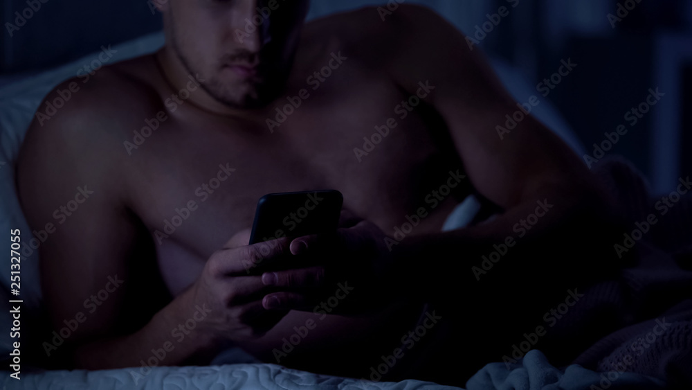 Topless man scrolling smartphone, lying in bed, checking e-mail before sleep Topless man scrolling smartphone, lying in bed, checking e-mail before sleep