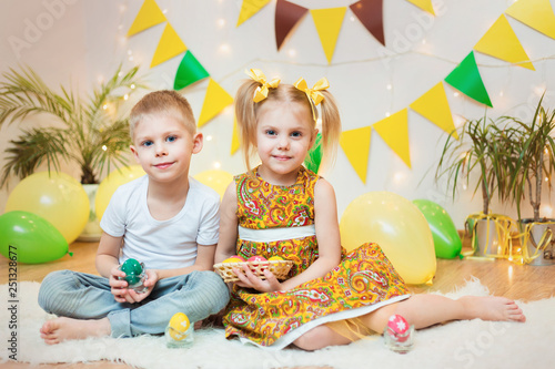 blond  blue-eyed cute smiling children boy in a white t-shirt and ponytail hair girl in yellow dress  brother and sister 4-5-6 years with Easter eggs in a room on the background of yellow decoration