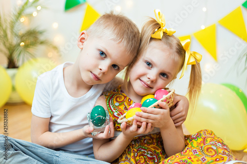 blond, blue-eyed cute smiling children,boy in a white t-shirt and ponytail hair girl in yellow dress, brother and sister 4-5-6 years with Easter eggs in a room on the background of yellow decoration