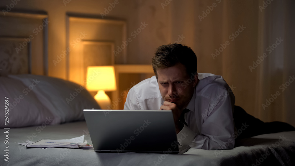 Exhausted engineer lying on bed, working on laptop at night, reading mail Exhausted engineer lying on bed, working on laptop at night, reading mail