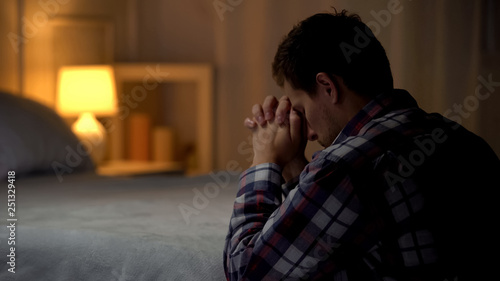 Religious young man praying in evening near bed, belief in God, Christianity