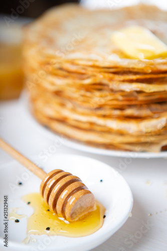 A stack of thin russian pancakes with butter and honey on a white plate