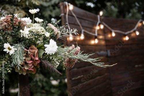 A flower bouquet at the foreground. A wooden photozone with a garland at the background.