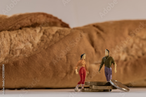 Miniature woman and man figure standing next to big baguette bread and counting coins. Shallow depth of field background. Family budget  healthy lifestyles and cost of food concept.