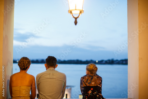 A bride, a groom and a bridesmaid are standing at the balcony and watching at the lake. photo