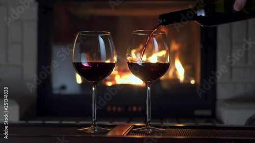 Male hand pouring red wine from bottle into two glass goblet flame of fireplace in background full shot. Romantic winter evening countryside atmosphere celebrating anniversary