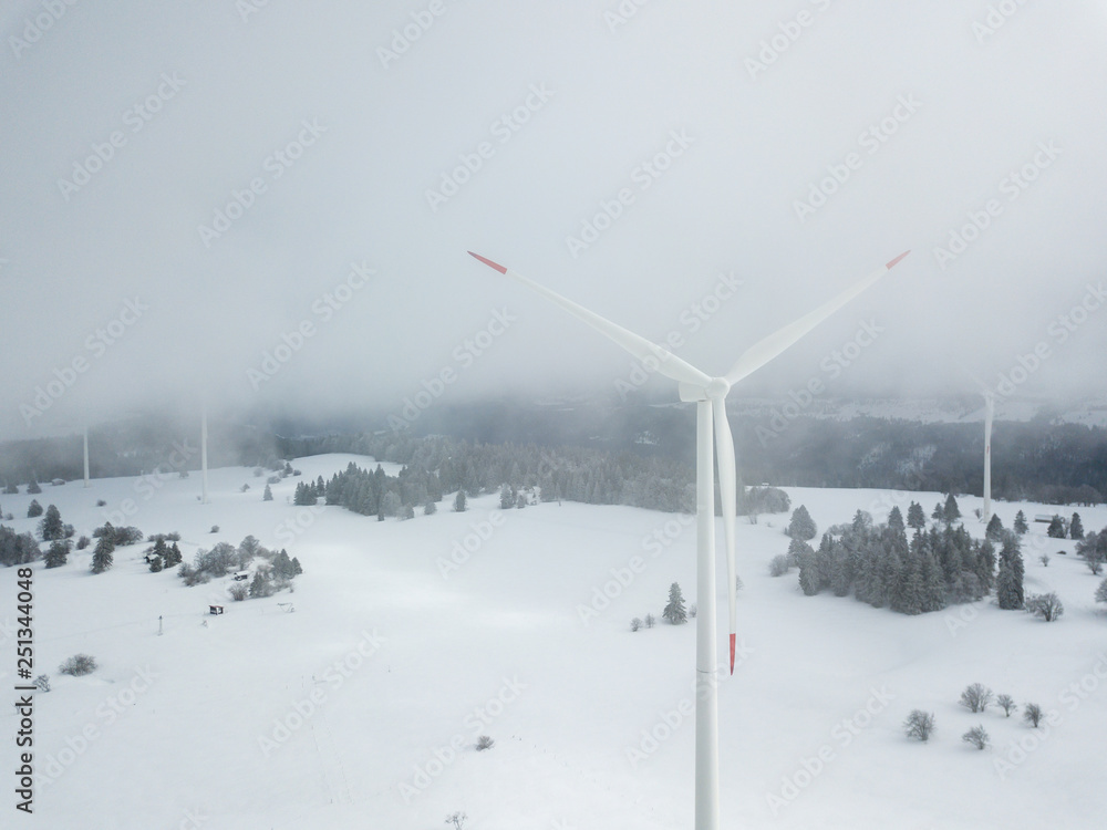 Aerial view of wind turbine in snow covered landscape in Swizterland. Tall pylon in fog, fir trees in the background. Aerial view of wind turbine in snow covered landscape in Swizterland. Tall pylon in fog, fir trees in the background.