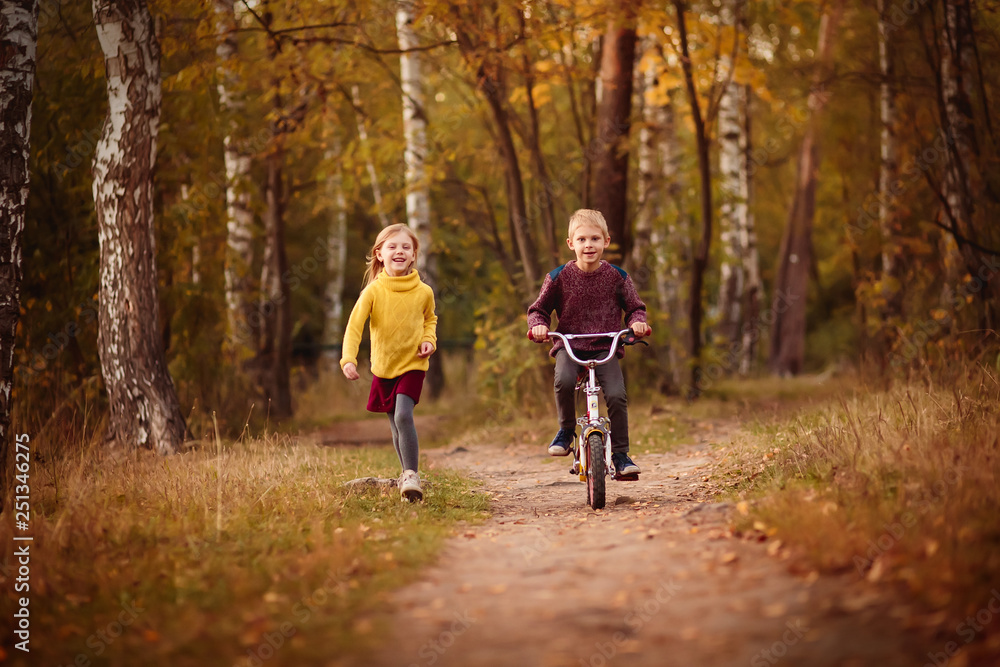 happy baby, baby boy 7 years riding a bicycle, and his little sister's sister, a little girl in a red hat runs with him along the park in the golden autumn happy baby, baby boy 7 years riding a bicycle, and his little sister's sister, a little girl in a red hat runs with him along the park in the golden autumn
