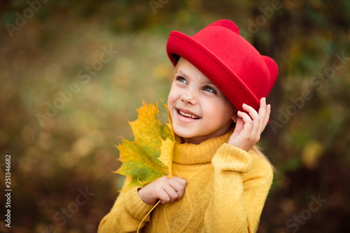 little cute happy little girl  6 year old baby in a red hat  a yellow sweater stands in a park or in a forest in the golden autumn and smiles