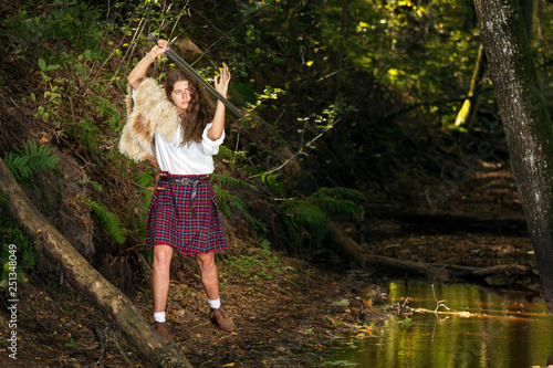 Girl in a Scottish kilt and animal skin on his shoulder posing in forest holding a Viking sword