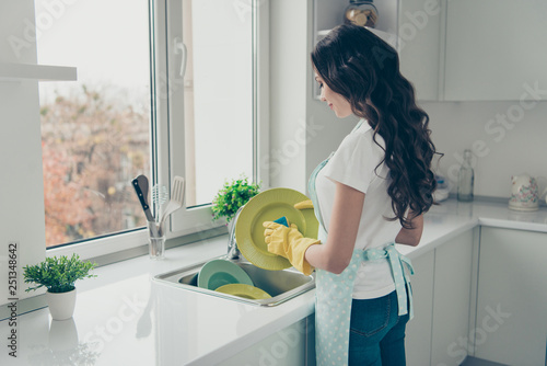 Profile side view portrait of her she nice charming lovely attractive beautiful cheerful wavy-haired house-wife washing green plates in yellow gloves in modern light white interior photo