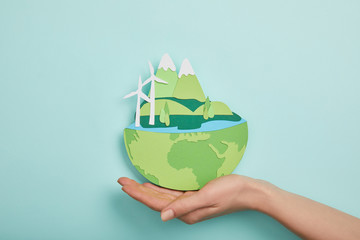 Top view of woman holding paper cut planet with renewable energy sources on turquoise background, earth day concept