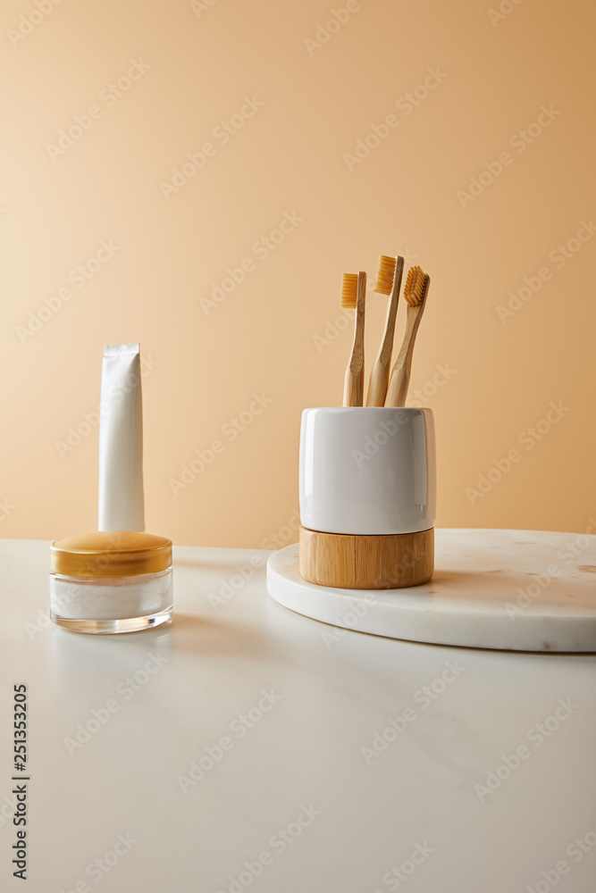 toothpaste in tube, cosmetic cream and holder with bamboo toothbrushes on white marble surface and beige background toothpaste in tube, cosmetic cream and holder with bamboo toothbrushes on white marble surface and beige background