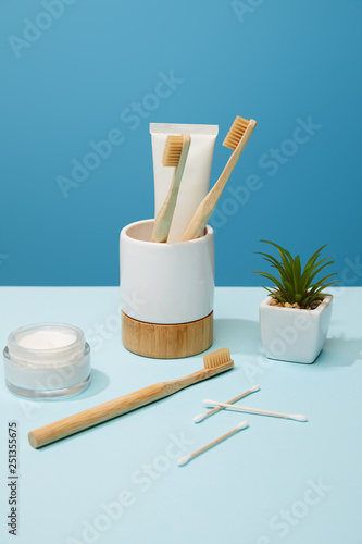 holder with toothpaste in tube and bamboo toothbrushes, cosmetic cream and plant in pot on table and blue background
