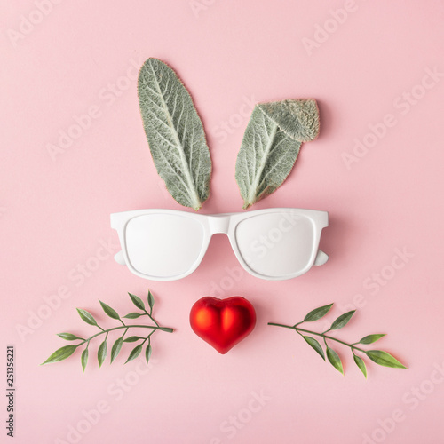 Happy Easter minimal concept. Bunny rabbit face made of natural green leaves with sunglasses and red heart on pastel pink background. Flat lay.