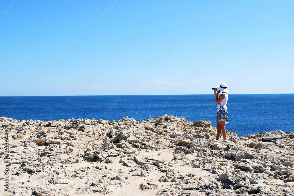 General view of the stony shore with a standing female figure in a white dress against the background of the sea, Kallithea, the island of Rhodes, Greece