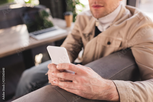 cropped view of trendy man sitting near laptop and paper cup and using smartphone