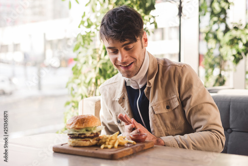 happy man looking at french fries near delicious burger on cutting board in cafe