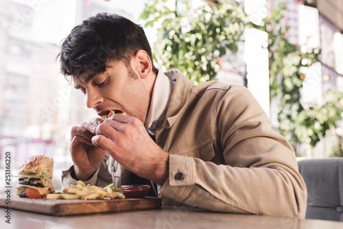 selective focus of young man eating tasty hamburger near french fries on cutting board in cafe