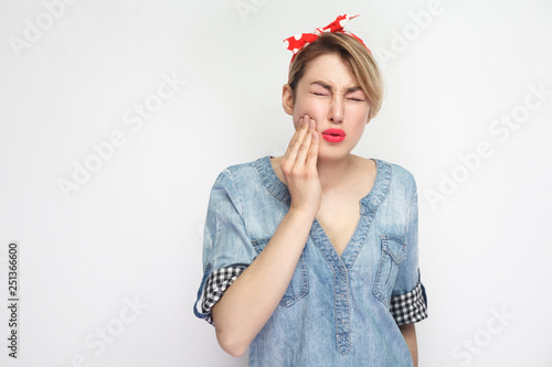 Tooth ache or pain. Portrait of young woman in casual blue denim shirt with makeup and red headband standing and feeling pain on her tooth. indoor studio shot, isolated on white background.