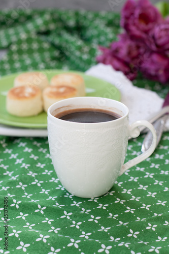 Black coffee in a white sophisticated cup, delicious dietary cheesecakes from home-made farmer cheese for breakfast, a bouquet of purple tulips on a green tablecloth. © myrka