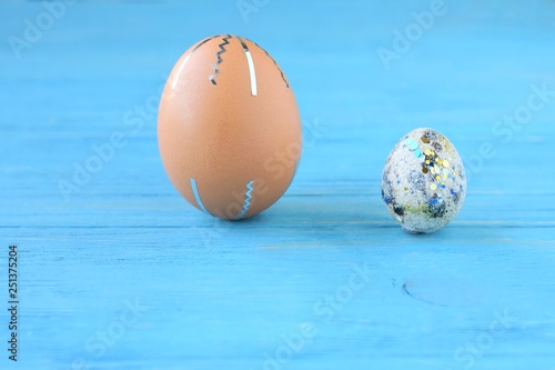 Brown chicken egg and quail spotted eggs with holiday Easter homemade decor on blue textured wood background with selective focus. Beautiful creative easter egg decor with blue shiny spots and stripes