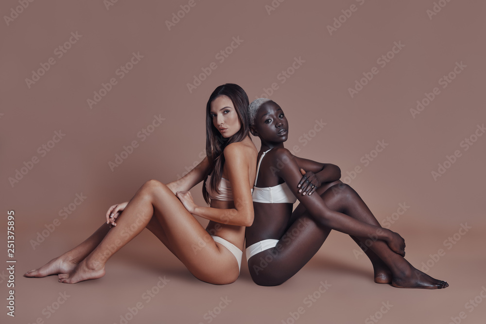 True feminine beauty. Two attractive mixed race women looking at camera while sitting back to back against brown background