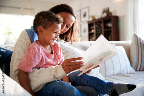 Close up of young mother sitting on a sofa in the living room reading a book with her toddler son, who is sitting on her knee, side view