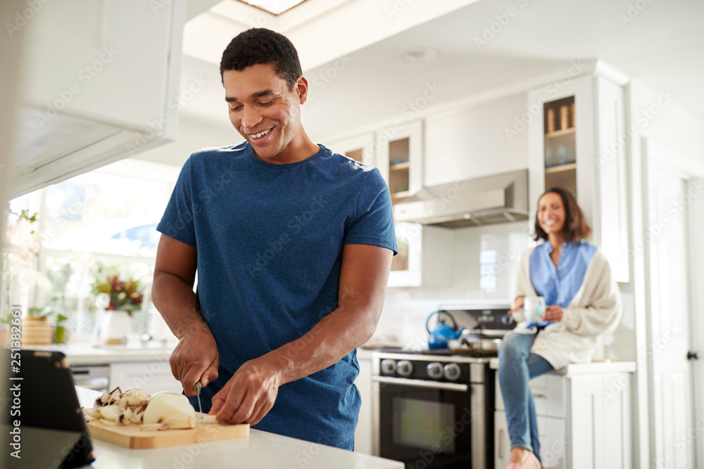 Young adult man standing in the kitchen preparing food, his partner sitting on kitchen worktop behind him, focus on foreground Young adult man standing in the kitchen preparing food, his partner sitting on kitchen worktop behind him, focus on foreground