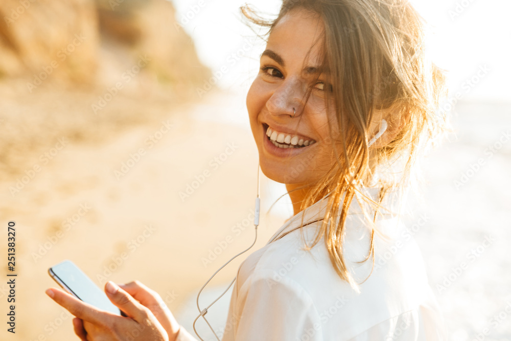 Beautiful young woman walking outdoors at the beach using mobile phone. - obrazy, fototapety, plakaty 