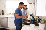 Young mixed race couple standing in the kitchen, man cooking food in a pan on the hob, close up, selective focus, side view