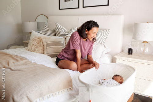 Young adult mother sitting on her bed looking down at her three month old baby sleeping in his cot photo