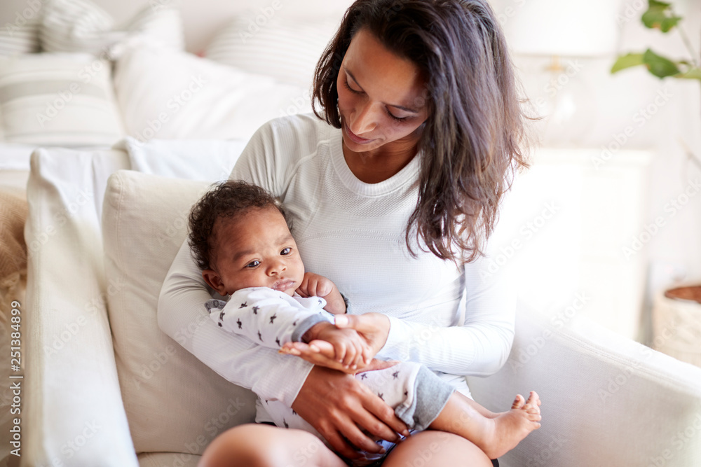 Young adult mother sitting in an armchair in her bedroom, holding her three month old baby son in her arms and looking down at him, close up Young adult mother sitting in an armchair in her bedroom, holding her three month old baby son in her arms and looking down at him, close up