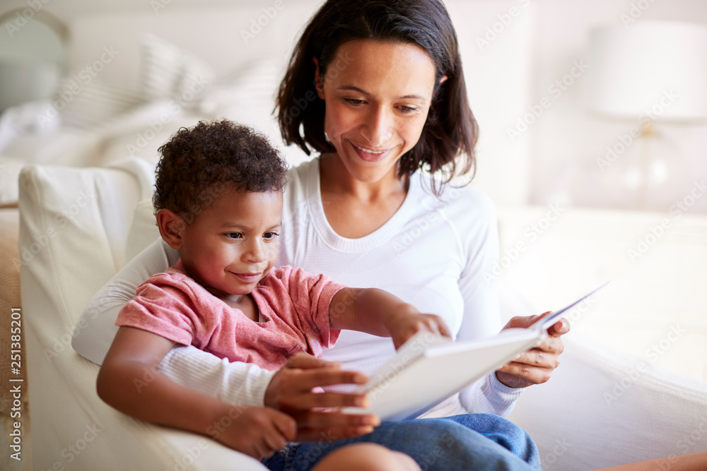 Close up of mixed race young adult mother sitting in an armchair reading a book with her three year old son on her knee, close up Close up of mixed race young adult mother sitting in an armchair reading a book with her three year old son on her knee, close up