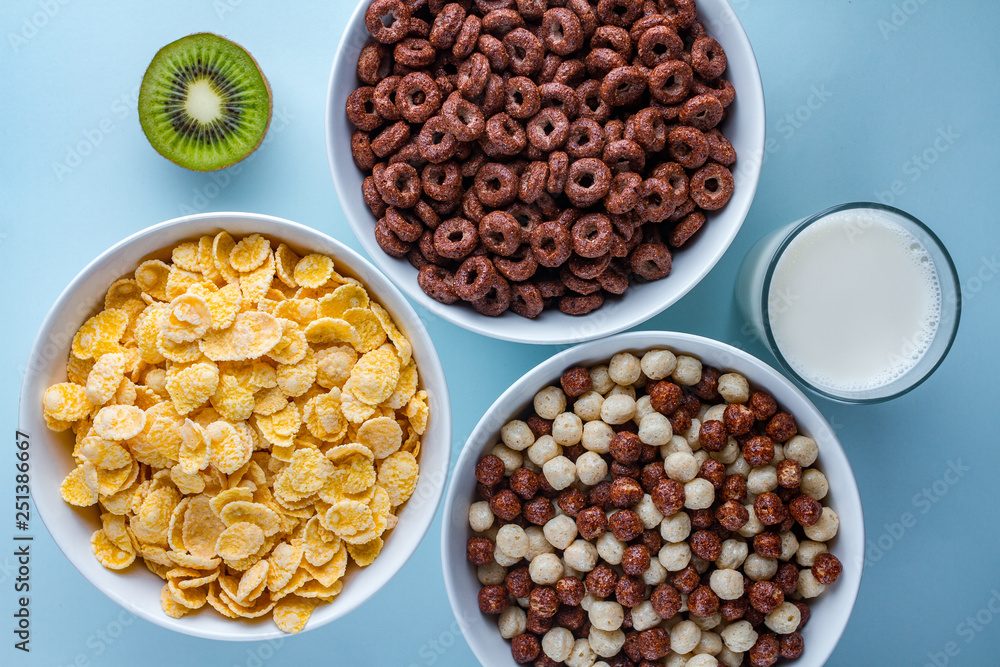 Cereals bowl of glazed, chocolate balls, rings, cornflakes, ripe kiwi and a glass of fresh milk for dry breakfast on blue background. Cereals bowl of glazed, chocolate balls, rings, cornflakes, ripe kiwi and a glass of fresh milk for dry breakfast on blue background.