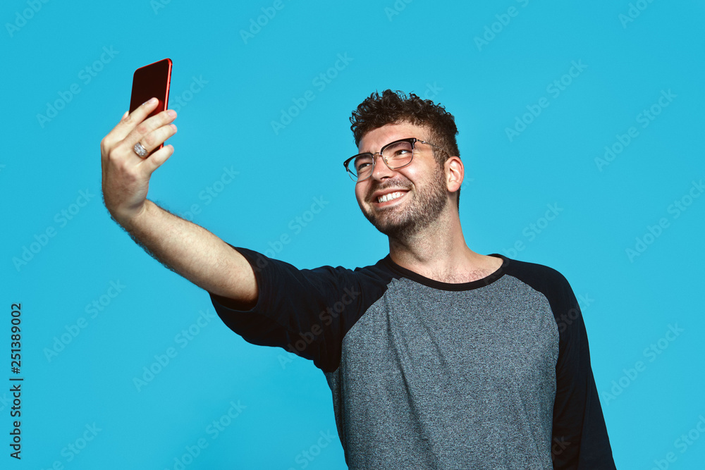 Cheerful hipster guy in trendy clothes using smart phone and taking selfie with smile, isolated over blue background Cheerful hipster guy in trendy clothes using smart phone and taking selfie with smile, isolated over blue background