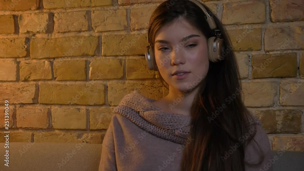 Young and beautiful caucasian girl in headphones sitting on sofa and watching calmly into camera on bricken wall background. Young and beautiful caucasian girl in headphones sitting on sofa and watching calmly into camera on bricken wall background.