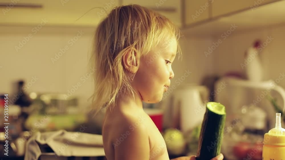 Stockvideo Naked child eating green cucumber in the kitchen. Little blonde girl holding a vegetable in her hands. | Adobe Stock